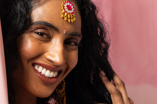 Indian woman smiling and touching hair while posing in studio with maang tikka, bindi, earrings