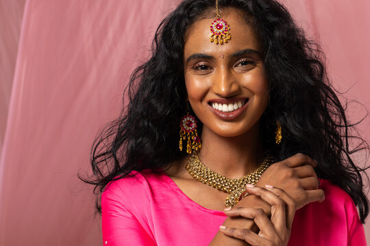 Indian woman smiling and holding hands near chest in studio wearing gold jewelry and pink dress
