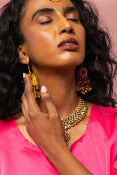 Indian woman tilting head in studio, touching gold earring with manicured hand in bright pink top