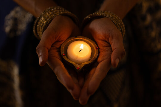 African American woman cupping lit candle in holder in dim room with gold bangles, metal trim