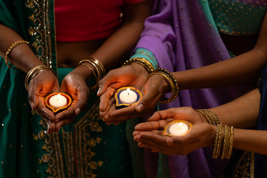 Indian women cupping and presenting lit clay diyas at home wearing ornate sarees and gold bangles