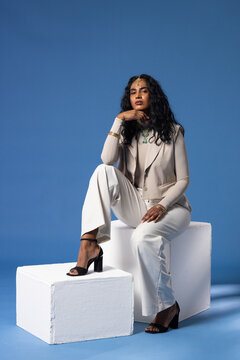 Adult female posing on white block at blue studio wearing beige blazer and turquoise necklace