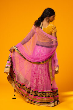 Indian adult woman posing, holding lehenga hem and draping dupatta on orange studio backdrop
