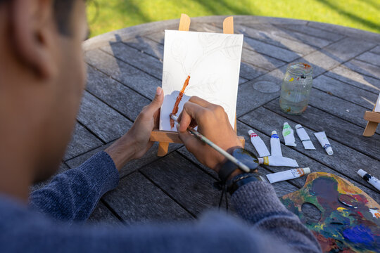 Indian man sitting at round wooden table in garden, painting leaf on canvas with paintbrush
