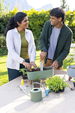 Diverse couple planting seedlings on lawn at backyard table using trowel and watering can