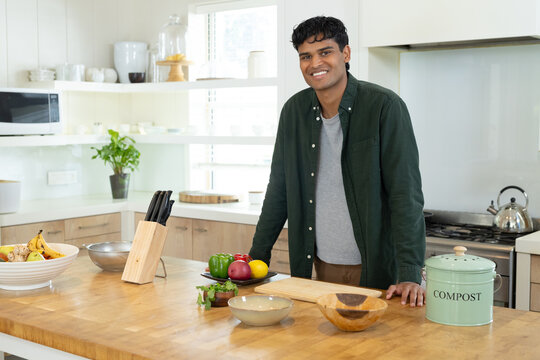 Indian man wearing green shirt and brown pants chopping at wooden island in kitchen near COMPOST