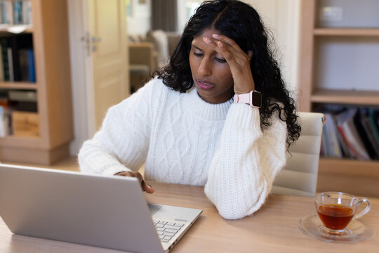 Woman wearing sweater rubbing forehead working on silver laptop at home desk with pink smartwatch