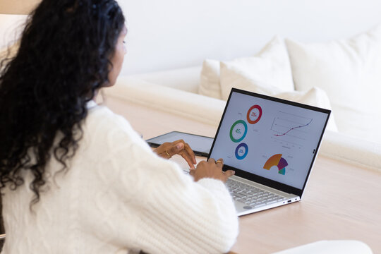 African American woman in sweater working on laptop with charts, tablet on wooden table at home