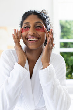 Female smiling placing red hydrogel patches under eyes wearing white bathrobe in bathroom by window
