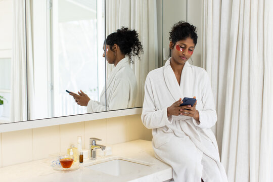African American woman sitting on vanity wearing robe, using phone, eye patches, mirror, copy space
