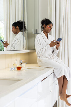 African American woman in bathrobe with patches sitting on bathroom vanity using phone, copy space