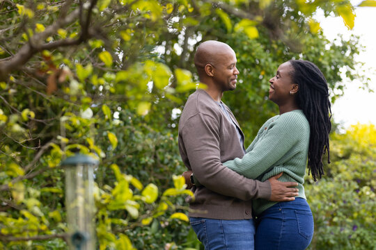 African American couple embracing, smiling in backyard by bird feeder, man wearing watch