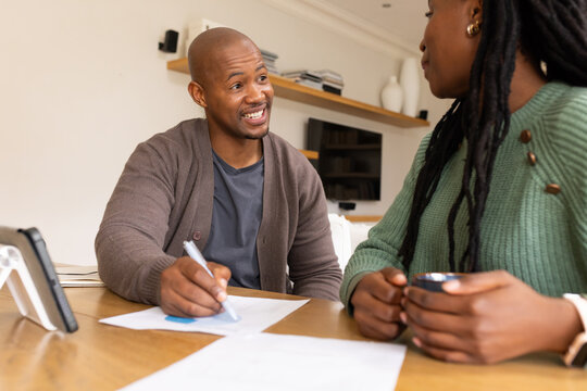 African couple reviewing papers at home table, man in cardigan holding pen, phone nearby