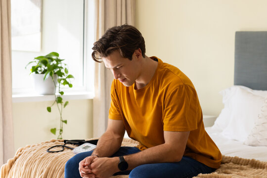 Adult man sitting on bed edge wearing mustard tee leaning forward, clasping hands beside BP monitor