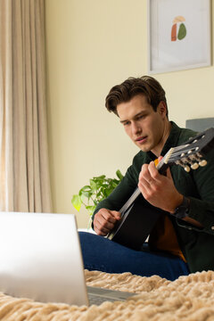 Man sitting on bed playing acoustic guitar while watching laptop in bright bedroom with plant