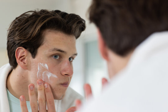 Man applying white facial cream with fingertips to left cheek at bathroom mirror, wearing bathrobe