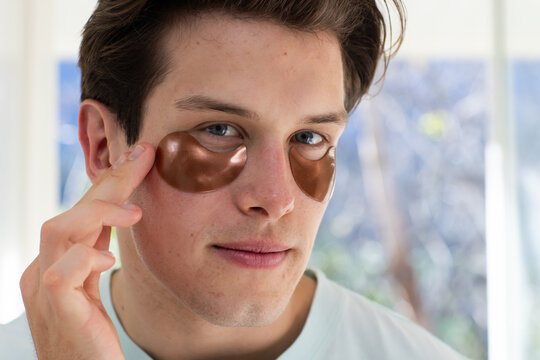Man applying metallic brown under eye gel patches near window, adjusting left patch with right hand
