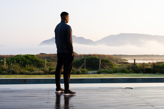 African American man standing on wet wooden deck beside pool in dark gear, facing misty mountains