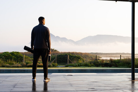 African American man wearing jacket on deck facing marsh carrying case and water bottle, copy space