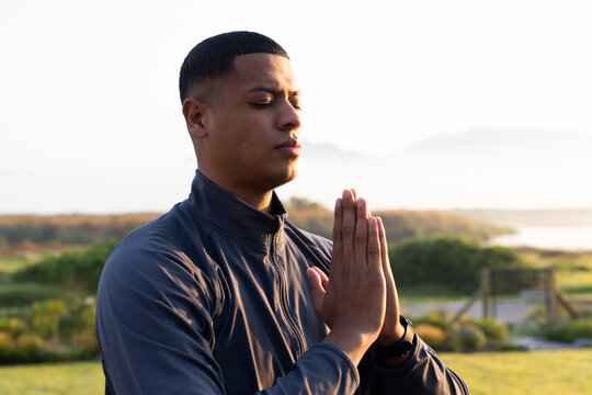 African American man standing praying on marsh path by wooden fence wearing zip-up jacket and watch