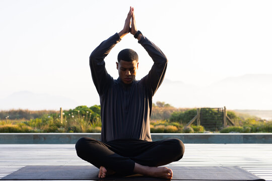 African American man in 20s cross-legged on deck with yoga mat raising palms above with smartwatch