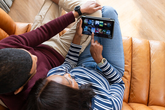 African American couple sitting on tan leather sofa sharing tablet, woman pointing media grid
