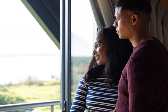 African American couple standing at window in living room, smiling and gazing at water, copy space
