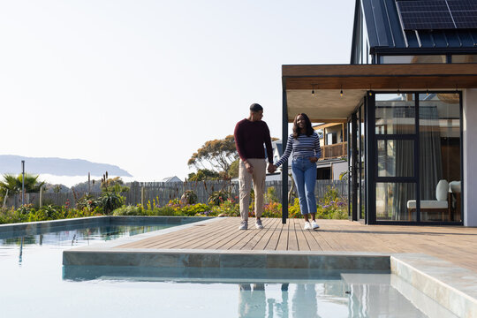 African American couple walking hand-in-hand on wooden deck by pool with modern house reflection