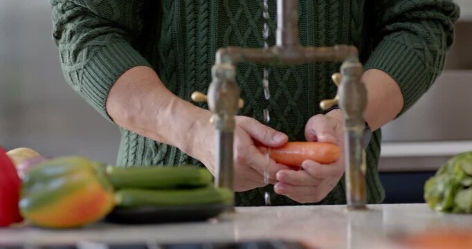 Asian man in sweater preparing carrots on cutting board and rinsing under faucet at kitchen counter