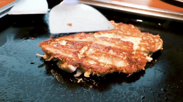 ヘラを使って鉄板の上でお好み焼きをひっくり返す調理シーン、香ばしい焼き目
