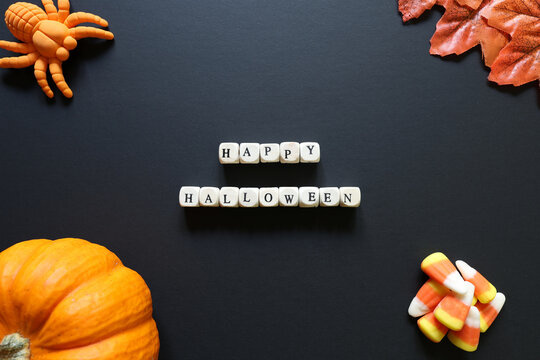 Happy Halloween message spelled with letter tiles on a dark background with pumpkin, candy corn, toy spider and autumn leaves.
