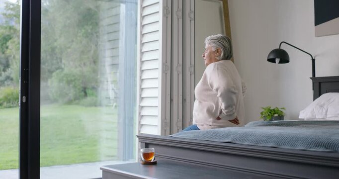 Senior African American lady sitting on bed rubbing neck for ease by glass sliding-door, copy space