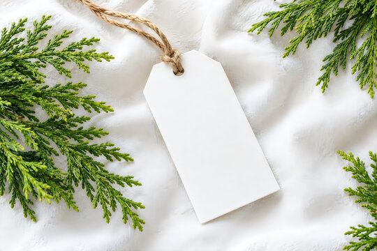 Elegant white gift tag with rustic twine placed on lush green evergreen branches set against a soft textured white linen fabric ideal for holiday or promotional designs