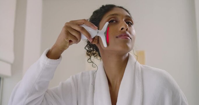 African American woman in robe touching jaw gliding T-redlight device on cheek at vanity for toning
