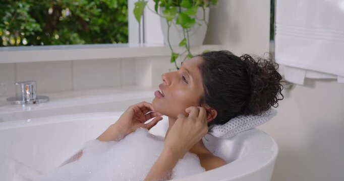 African American woman reclining in bubble bath by window placing earbud and closing eyes for music