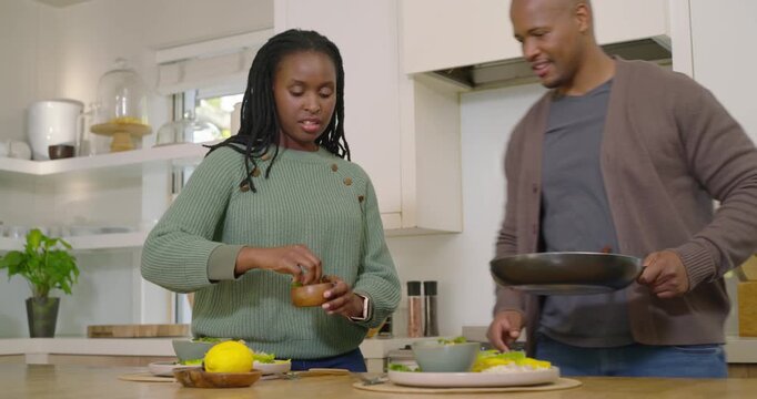 African American couple preparing at island, man holding pan, mixing greens, grinding pepper