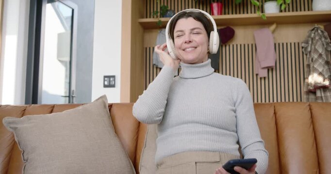 Adult woman in gray sweater tapping smartphone, placing white headphones and queuing music on couch