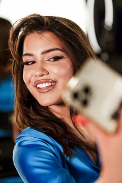 Attractive smiling female capturing a self portrait using mobile phone camera during professional photoshoot, emphasizing cosmetic makeup and trendy style.