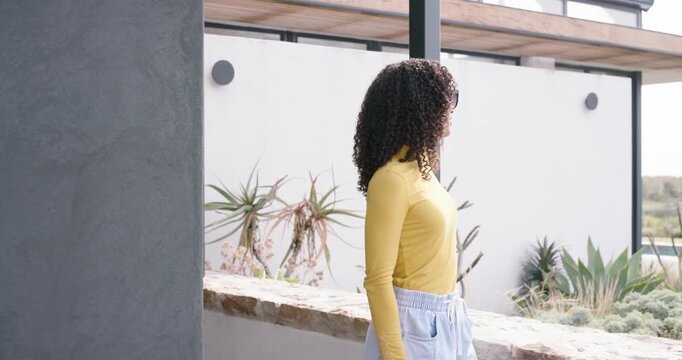 African American woman in yellow top picking up sunglasses stepping onto patio ledge scanning view