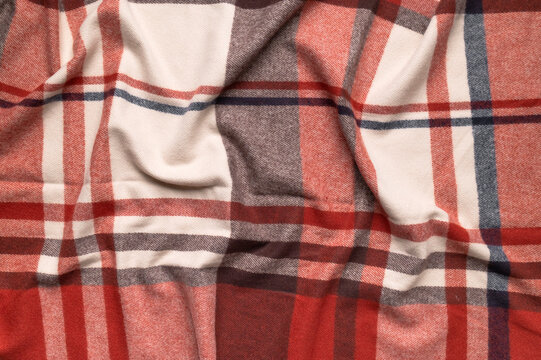 Warm plaid fabric forms soft waves in close view, showing red, beige and gray checks with tactile weave. Textile material suits scarf design, autumn clothing, cozy styling and retail use.
