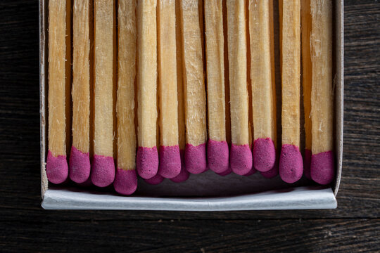 Matches with pink heads in cardboard box closeup. Top view of wooden matches with purple sulfur heads inside a matchbox. Row of matchsticks on dark background for conceptual design or marketing