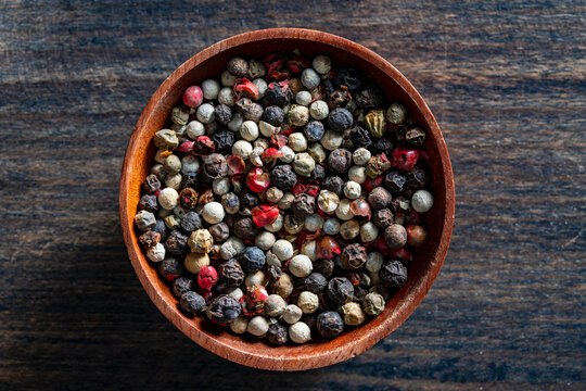 Peppercorn mix in wooden bowl closeup top view. Top view of colorful pepper blend in small wood bowl. Mix of black white green and pink peppercorns on dark wooden background for culinary design
