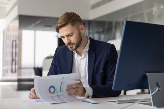 Male marketing specialist engaged in paperwork at office desk reviewing printed business reports reading paper documents with charts and diagrams checking sales statistics studying project analytics