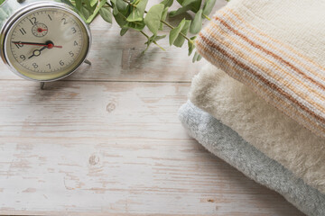 An alarm clock and stacked clean towels with eucalyptus on white wooden background with copy space for morning routine concept. © PH_BRICK