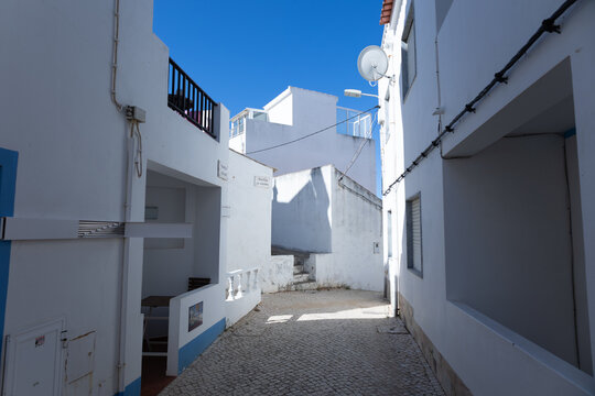 Narrow whitewashed cobblestone alley with blue accents under a clear blue sky in Burgau, Algarve, Portugal