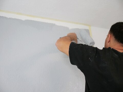 Professional painter using a paint roller to apply grey color on interior wall near the ceiling during room renovation and home improvement work.