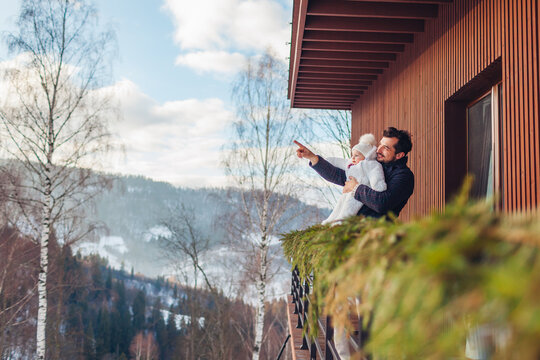 Dad and his baby girl enjoy snowy mountain landscape from hotel terrace. Family spends weekend in rented house for winter holidays