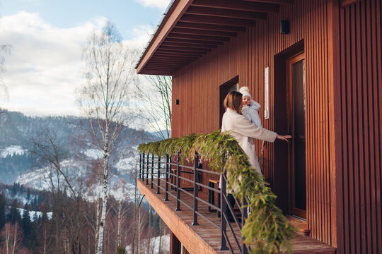 Happy family renting cabin in snowy mountains for Christmas. Woman enters house decorated for holidays holding toddler baby girl