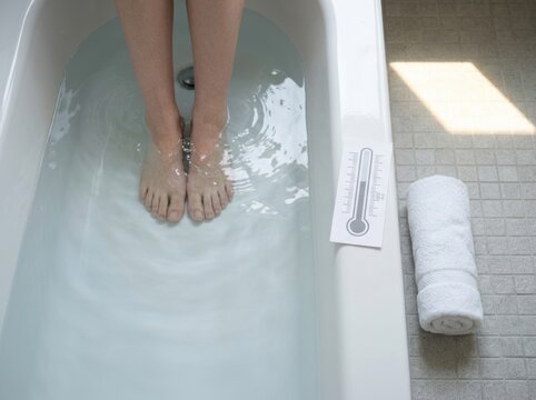 Bare feet of a person submerged in a bathtub filled with water, thermometer and towel nearby