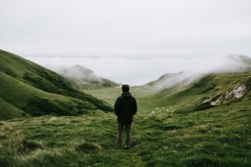 Fototapeta premium A lone figure stands on a cliffside at dawn overlooking a distant valley and mountains while clouds slowly drift across the landscape below